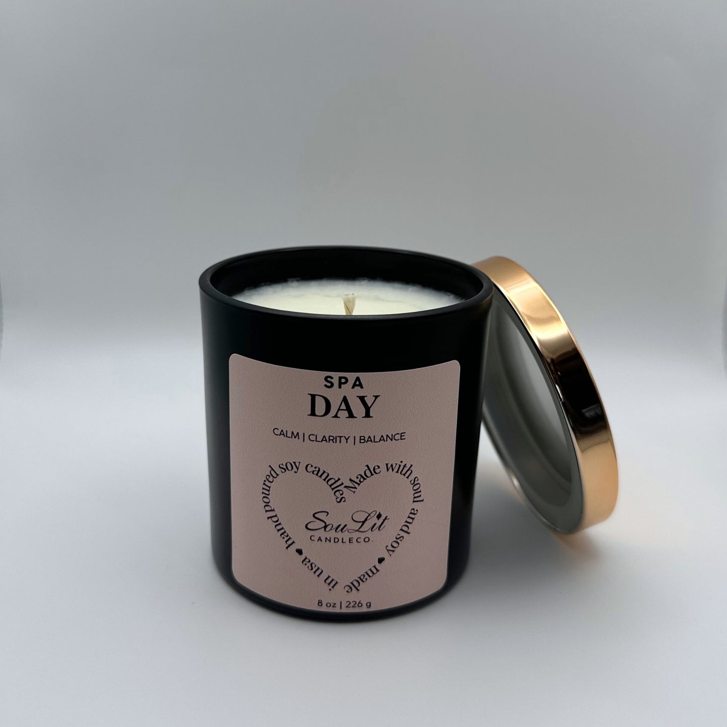 Soy candle in 8oz matte black glass jar with rose gold lid off propped on the side of jar on white background. 