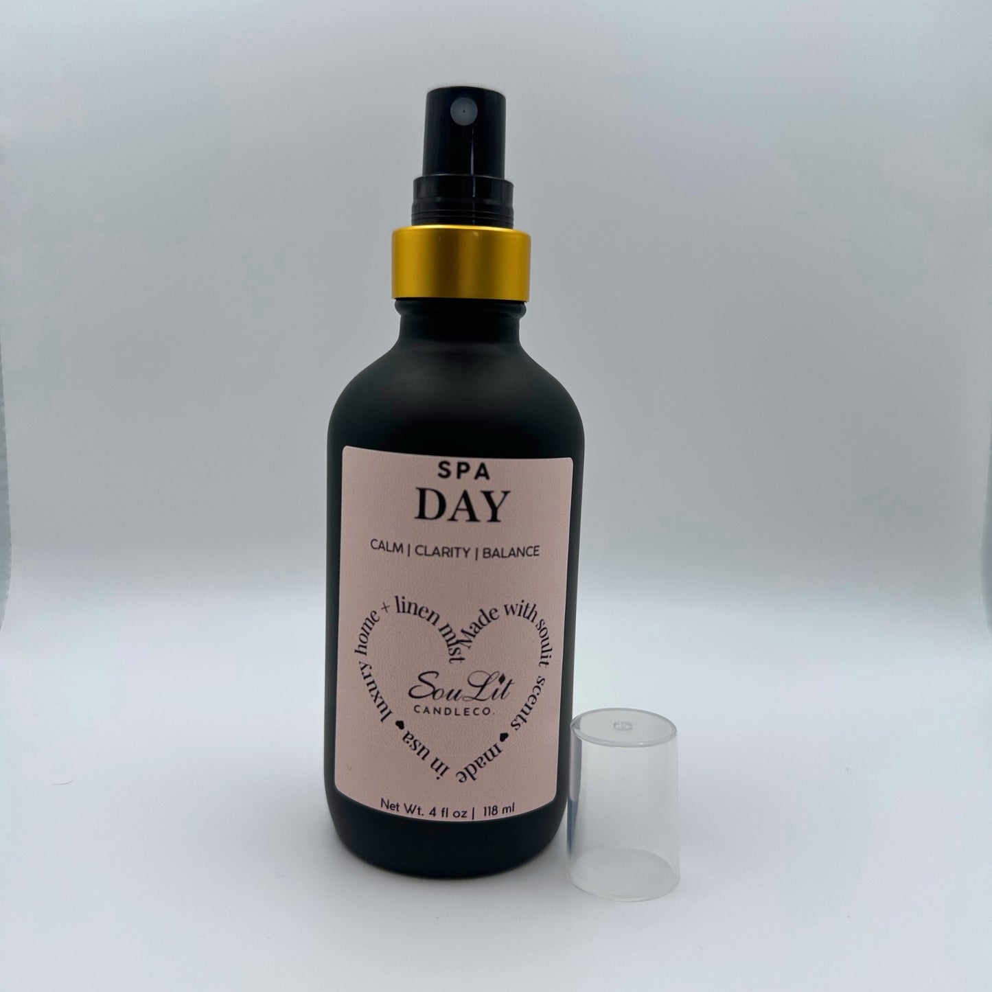 Air and linen freshener mist in 4oz matte black glass bottle with a gold neck with clear top on the side on white background. 