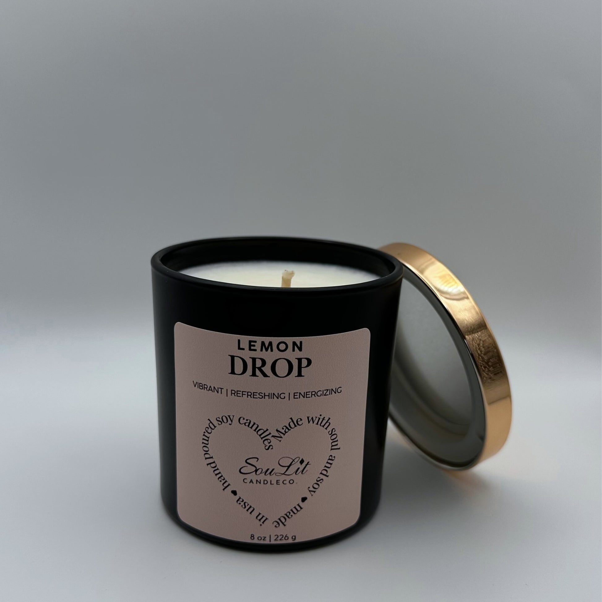 Soy candle in 8oz matte black glass jar with rose gold lid off propped on the side of jar on white background. 