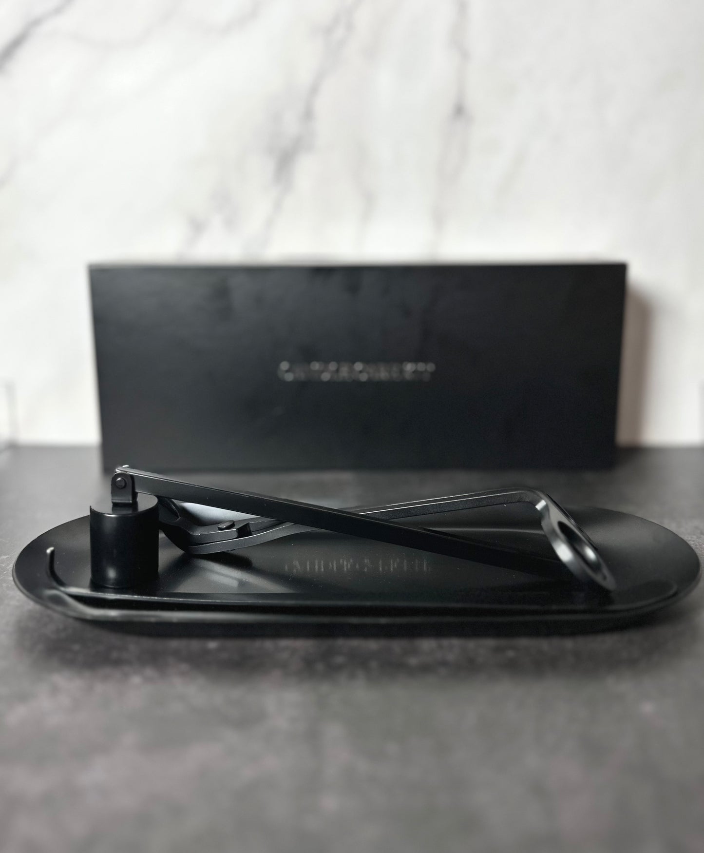 Side view of the semi-gloss black 4-piece candle care kit with wick trimmer, snuffer, and dipper on tray with the matte black product box blurred in background.