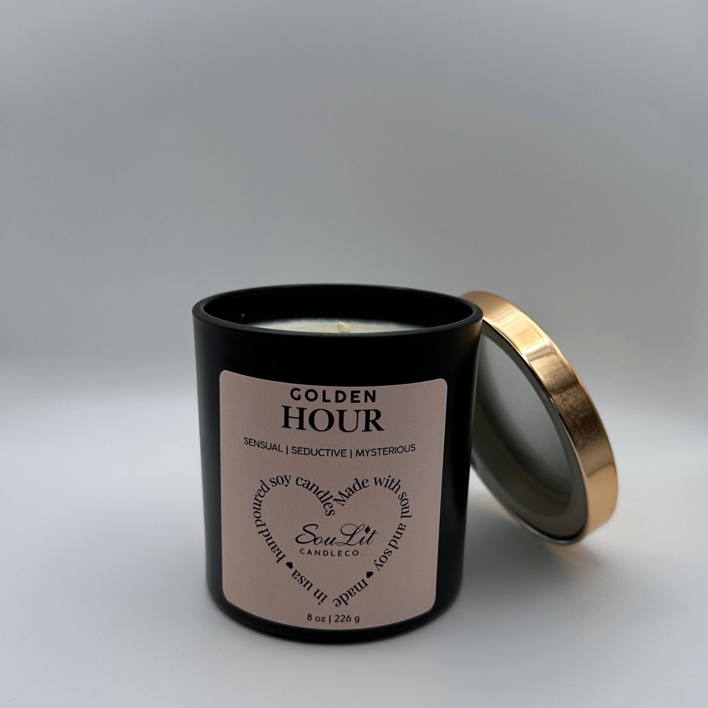 Soy candle in 8oz matte black glass jar with rose gold lid off propped on the side of jar on white background. 