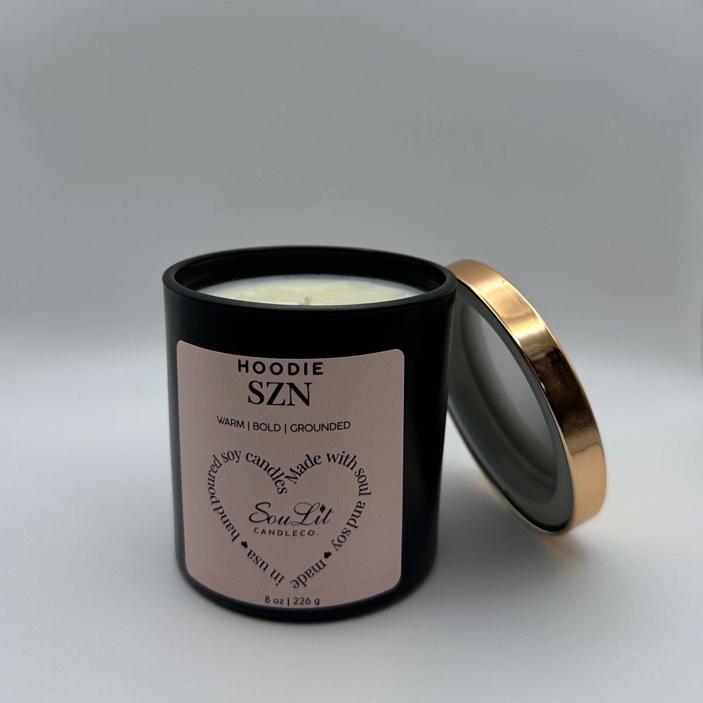 Soy candle in 8oz matte black glass jar with rose gold lid off propped on the side of jar on white background. 