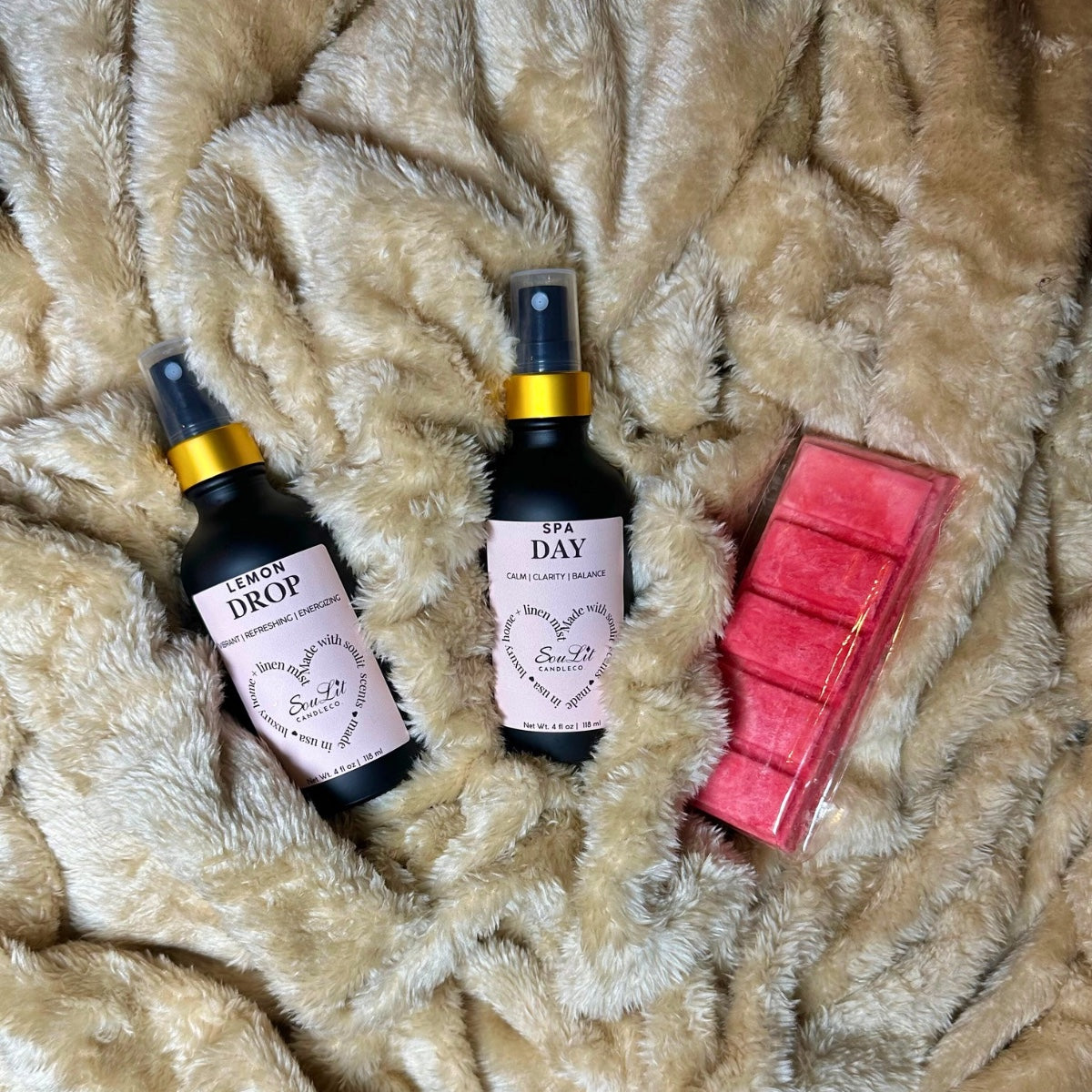 Two air and linen sprays in 4oz matte black glass bottles with gold neck and a wax melt bar on a textured beige surface.