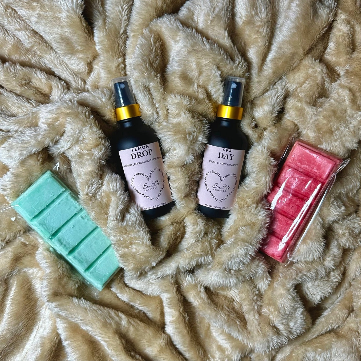 Two air and linen sprays in 4oz matte black glass bottles with gold neck and two wax melt bars on a textured beige surface.